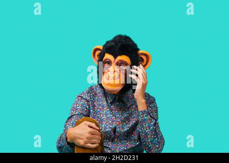closeup of a young man wearing a monkey mask talking on the phone on a blue background Stock Photo