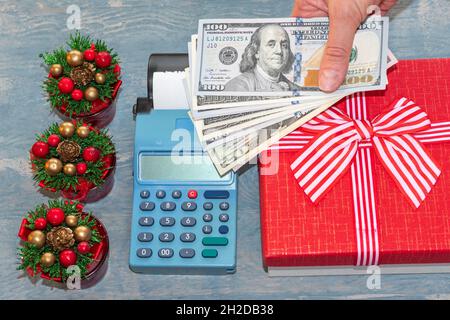 Christmas sale. A hand holding dollars over a cash register near a red gift box with a ribbon and small Christmas trees with cones. online shopping. B Stock Photo