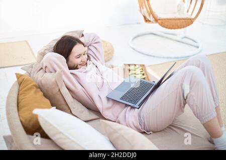 Young smiling woman works on the laptop lying on sofa Stock Photo