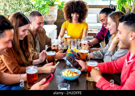 young multiethnic group of friends holding smartphone on social media having fun together drinking happy hour beer at bar restaurant sitting on table Stock Photo