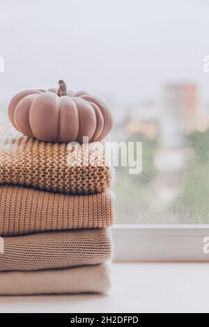 A stack of warm knitted sweaters on the windowsill with pumpkin on a rainy day, in pastel shades. Stock Photo