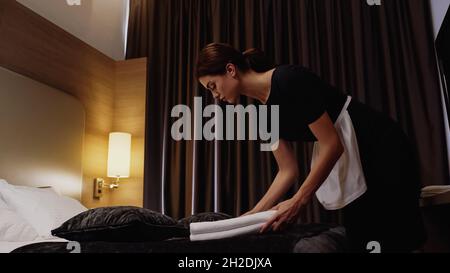 young maid putting fresh white towels on bed in hotel room Stock Photo