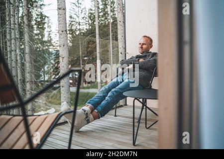 Portrait of a Middle-aged man dressed open cardigan, jeans, and warm socks sitting on forest house balcony and enjoying the fresh air. Everyday lifest Stock Photo