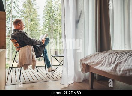 Alone man sitting in chair on country house balcony and enjoying forest view, tea cup, fresh air and bestseller novel thick book. Cozy bedroom interio Stock Photo