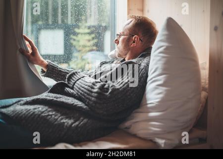 Portrait of thinking sad Middle-aged man in eyeglasses dressed open cardigan lying on cozy bed next to window looking out street through raindrop glas Stock Photo