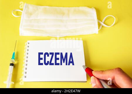 The phrase Eczema with on a notebook is written by a female hand. On a yellow background, a syringe and a medical mask Stock Photo