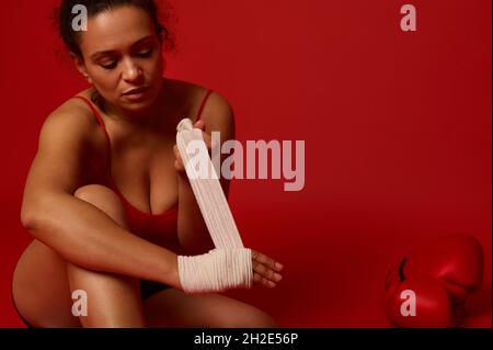 Confident Afro American female fighter, boxer sitting on colored background and tying hand wrap, preparing for the competition waving her hands with a Stock Photo