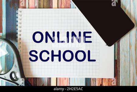 ONLINE SCHOOL - Text written in a notebook next to a mobile phone and a magnifying glass on a striped table. Flat composition. Back to school, start o Stock Photo