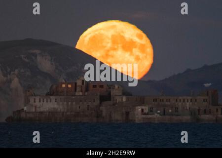Portland, Dorset, UK.  21st October 2021.  UK Weather.  The almost full Hunter’s Moon rises up from behind the cliffs of the Jurassic Coast above the historic 19th century Portland Breakwater Fort on the outer breakwater of Portland Harbour near Weymouth in Dorset.  The now derelict fort also known as Chequered Fort was constructed between 1868 and 1878 and is Grade II listed.  Picture Credit: Graham Hunt/Alamy Live News Stock Photo