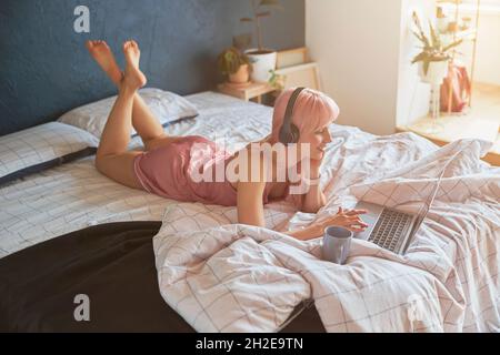 Smiling lady with headphones works on laptop lying on double bed Stock Photo