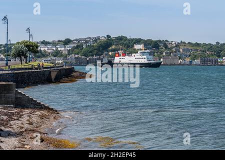 Calmac ferry leaving Rothesay harbour en-route to Wemyss Bay seen from esplanade  with sea shore and seaweed in foreground and town in background. Stock Photo