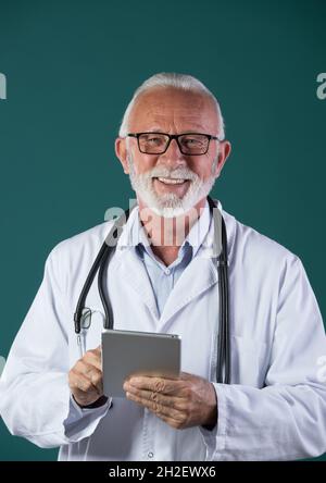 Portrait of senior doctor wearing white coat, with stethoscope around neck, holding tablet, smiling and looking at camera, against blue background Stock Photo