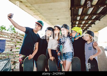 large group friends children skateboarders make videos, online broadcasts, video calls on phone in skate park. Kids spend time on skateboarding traini Stock Photo