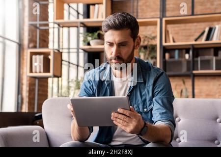 Serious caucasian man using digital tablet sitting on sofa at home office Stock Photo
