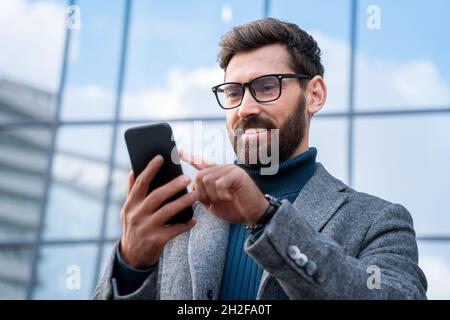 Smiling businessman using smartphone standing outside office in urban street Stock Photo