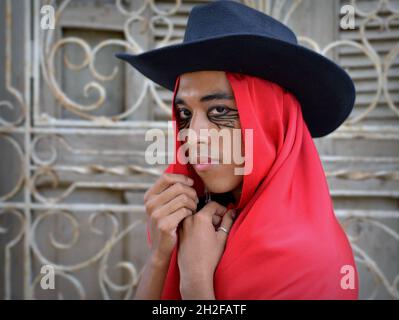 Handsome young nonbinary Latino wears a black hat with red Mexican rebozo as a hood and poses in front of a white wrought iron window grill. Stock Photo