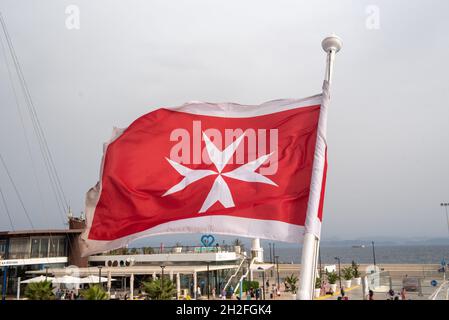 Maltese flag on a luxury boat anchored in the marina of Greece. Old Maltese flag, white cross on red background waving at stern of yacht. Blue sky bac Stock Photo