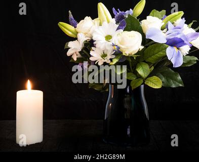 Funeral Bouquet purple White flowers and burning white candle, Sympathy and Condolence Concept on black background with copy space. Stock Photo