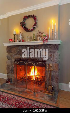Traditional fieldstone wood burning fireplace with a cosy fire and mantlepiece with Christmas decorations and a holiday wreath. Stock Photo