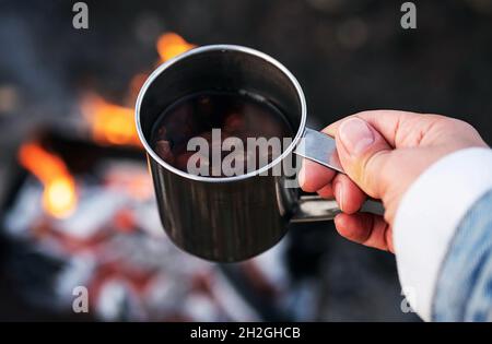 Woman hand hold rose hip tea in metal mug against bonfire. Eco-friendly tourism, sustainable lifestyle. Stock Photo