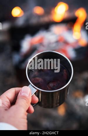 Woman hand hold rose hip tea in metal mug against bonfire. Eco-friendly tourism, sustainable lifestyle. Stock Photo