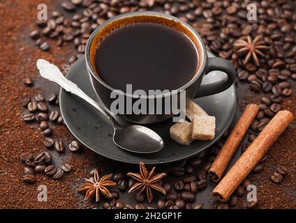 Black coffee in ceramic cup with cinnamon and cane sugar with anise star and silver spoon on ground coffee and beans background. Stock Photo