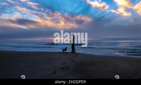 Silhouette of a man and his dog standing together and facing each other at the beach with a beautiful dramatic sunset sky in the background Stock Photo