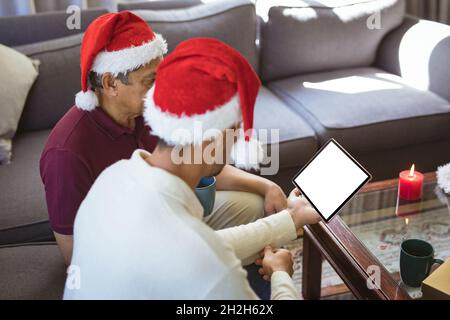 Biracial adult son and senior father in santa hats making tablet christmas video call Stock Photo