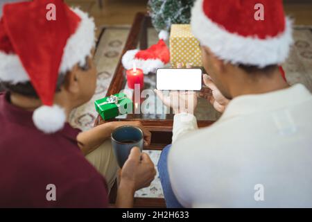 Biracial adult son and senior father in santa hats making smartphone christmas video call Stock Photo