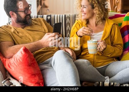 Happy couple sitting on sofa and looking at each other while drinking coffee and using mobile phone at home. Loving couple spending leisure time toget Stock Photo