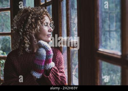 Adult pretty woman looking outside the window at home in winter season. Thoughtful happy female people enjoy cold season using gloves at home and drea Stock Photo