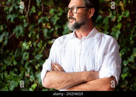 Thoughtful mature man wearing eyeglasses and looking away while standing with his arms crossed in front of green leaves background. Bearded man lookin Stock Photo