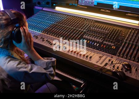 Rear view of young woman listening to music in headphones while using music keyboard in the recording studio Stock Photo