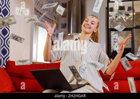 The girl made a lot of money online from home. Girl tossing hundred dollar bills while sitting on sofa with laptop Stock Photo