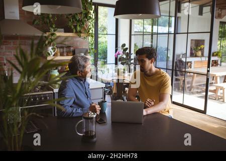 Biracial adult son and senior father talking and using laptop in kitchen Stock Photo