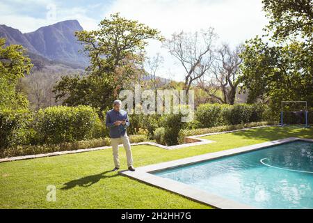 Senior biracial man using smartphone at swimming pool in garden Stock Photo