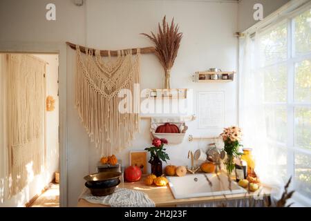 Cozy rustic kitchen with large window and lace curtains. On the wall hangs a large macrame, pumpkins, cereals Stock Photo