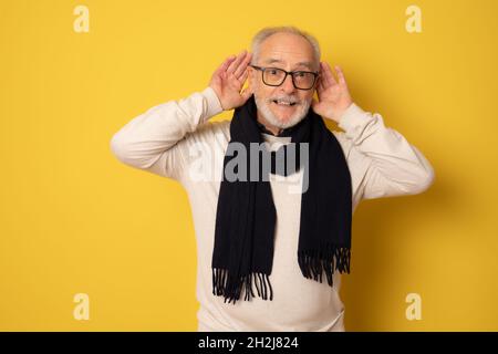 Senior man wearing winter clothing trying to hear both hands on ear gesture, curious for gossip. Hearing problem, deaf. Stock Photo