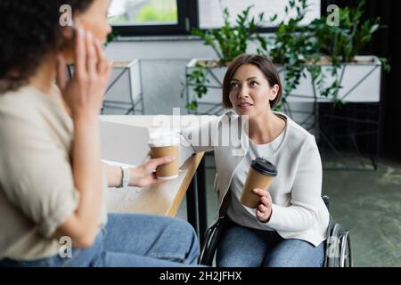 businesswoman in wheelchair talking to blurred african american colleague sitting on desk during coffee break Stock Photo