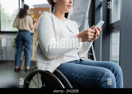cropped view of businesswoman in wheelchair messaging on mobile phone near african american colleague on blurred background Stock Photo