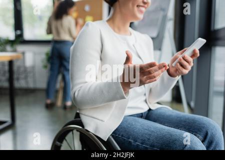 cropped view of cheerful businesswoman in wheelchair gesturing while using mobile phone near blurred african american colleague Stock Photo
