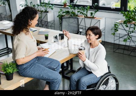 african american businesswoman sitting on desk during coffee break near colleague in wheelchair Stock Photo