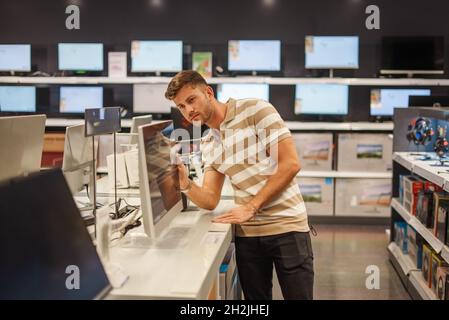 Man in casual clothes looking closely at screen of modern monitor placed on shelf in electronics department of mall Stock Photo