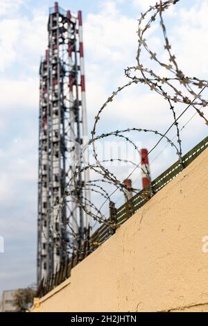 barbwire on yellow fence and factory pipes and blue sky with white clouds on background (focus on the wires) Stock Photo