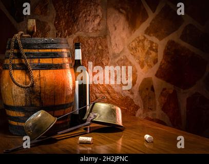 Wine Mood. A barrel and two wine ladles on a table against a stone wall. In the center of the composition is a black bottle with a white label. Stock Photo