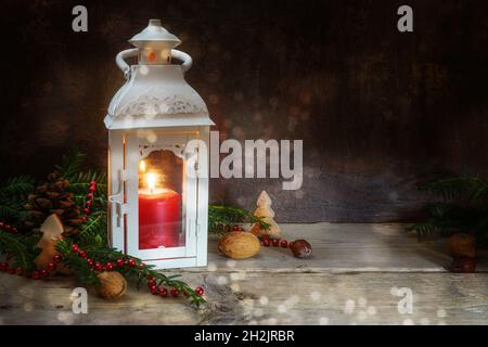 Christmas and Advent decoration, red candle with warm light in a white lantern, fir branches and nuts on rustic wooden planks against a dark bokeh bac Stock Photo