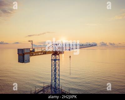 Close-up view from a drone of a construction tower crane against the background of a fantastic, beautiful sunset on the sea with a copy of the space Stock Photo
