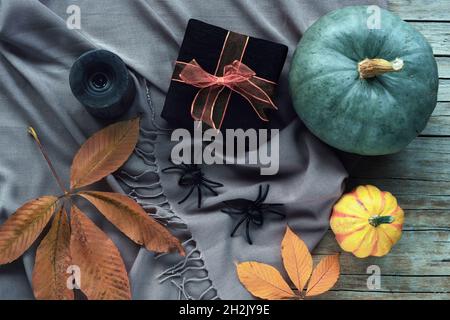 Pumpkins, yellow leaves, gift box in black wrapping paper, spiders and candles on gray scarf background on wooden table. Halloween concept. Top view, Stock Photo