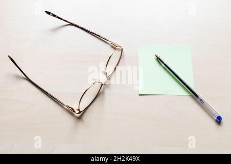 top view of eyeglasses and simple blue pen on green sheet of note paper on light brown wooden board Stock Photo
