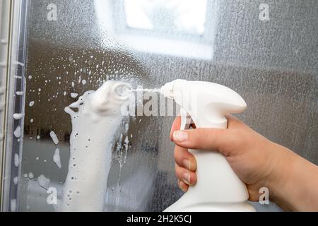 Person hand using limescale remover chemical foam. Descaling shower case glass door. Work in progress. Stock Photo